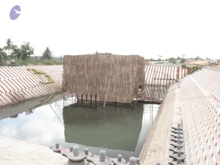 Photograph of an outdoor construction site at a water-filled rectangular tank with sloped stepped sides faced with red and white striped tiles. In the centre a large square structure is clad with stacked grey-brown wooden planks or bamboo-like sheathing, standing on a scaffold frame above the water and reflected in it. Several grey stone shivalingam-style pedestals with round tops line the lower edge of the near steps. Palm trees, shrubs and a clear pale sky sit beyond, with construction machinery and piled materials visible at the far right. No people are visible.