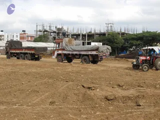 A photograph of a dirt construction site under a cloudy sky. Two large trucks with tarpaulin-covered cargo beds, one red and white, drive across unpaved ground, and a red Massey Ferguson-style tractor with a canopy sits at right. Behind them stands a partly built multi-storey concrete frame with exposed steel reinforcement rods rising from the columns, and a pile of rebar lies beside the trees. A small figure stands on the upper level of the frame. Low white and red buildings and green trees line the background. No people are clearly visible besides a worker on the structure and tractor and truck drivers.