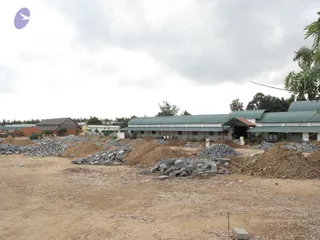 A photograph of a dusty construction site at an ashram campus. Piles of cut grey stone slabs and mounds of reddish-brown earth lie scattered across bare ground in the foreground. Behind them stands a long white single-storey building with green corrugated metal roofs and a covered entrance porch. A few small figures in white clothing stand near the building, and a tall tree and low brick structures are visible at left and right under an overcast sky.