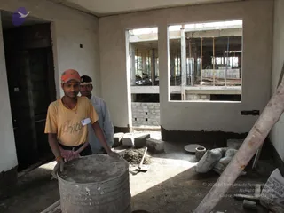 Two young male construction workers stand inside an unfinished concrete building with plastered white walls and an open window frame. The foreground worker wears an orange cap, a yellow t-shirt, a red-and-white checked lungi tied at the waist, an orange wrist thread and a beaded necklace, and he rests his hands on a grey cement drum. A second worker behind him wears a white shirt and a white headband. Nearby on the concrete floor are stacked cement blocks, a trowel, a heap of mortar, cement sacks and a rusty steel pipe leaning against the wall. Through the window, a multi-storey concrete frame with bamboo or steel supports and stacked brick walls is visible.