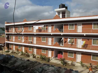 A photograph of a newly built three-storey brick residential block with white-trimmed balconies, metal railings and a water tank on the roof, at the Bidadi Ashram construction site. Laundry hangs from several balconies, potted plants line the ground floor entrances, and corrugated metal sheets with cut tree branches lie in the foreground. A single figure in an orange-saffron robe walks along the lane at the lower right edge. Electrical cable runs diagonally across the upper left.