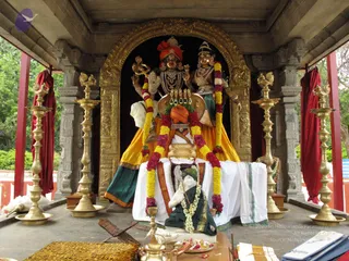 A temple shrine under a stone pavilion roof with a gold-framed arch backdrop. A large deity murti with a red and orange turban, gold and white ornaments and a trident-style staff stands beside a consort figure in green and yellow dress, both garlanded with yellow and magenta flower strands. In front sits a white-draped peetha with a small white and green robed figure with a garland, tall brass oil lamps flank the shrine, and brass puja vessels, a plate of flowers and a tiger-striped cloth sit on the floor. No living people are visible.