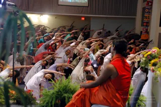 A large group of participants in a hall performs a standing side-stretch yoga exercise, arms raised overhead and bodies leaning to one side in rows. Most wear white, cream, lavender and blue clothing. In the foreground on the right, a person in a saffron-orange robe sits with back partly turned to the camera, leading the session from a low seat beside colourful flower arrangements. Green ferns and a leafy plant frame the left edge, and a projection screen and wall banner appear at the top.