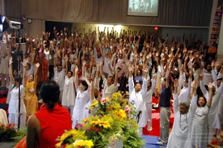 A photograph of a large group session in a hall, with a crowd of several hundred people standing with both arms raised overhead, palms open. Most wear white or light clothing; a few wear red, yellow and orange. In the foreground at the lower left, a woman in a red sleeveless top faces the group, and a bouquet of yellow sunflowers, pink roses and greenery sits on a table in front of a microphone. A stage light stands at the left edge, and a projection screen hangs at the top. Red mats cover the floor. Near the left, a figure in orange robes stands among the crowd.