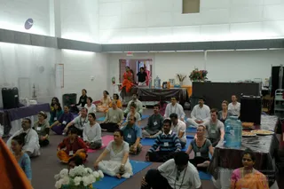 A large indoor hall with white walls and bright ceiling lights hosts a seated group of about 30 yoga participants on blue, pink and purple mats. Most sit cross-legged facing forward, some in white kurtas, others in T-shirts, striped sweaters or patterned tops, many wearing name badges on lanyards. At the front, a woman in an orange sari stands near a draped table holding a small arrangement, with a bouquet of flowers, a water cooler and platters of food on side tables. Two people stand near the doorway at the back. A white bouquet sits in the foreground.