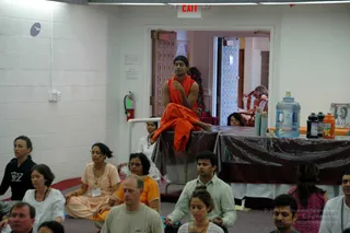 A photograph of a group meditation or satsang in a bright indoor hall with white cinderblock walls and a red EXIT sign. A man in orange robes and an orange shawl sits cross-legged on a draped table at the back, a candid appearance-based candidate for a person (unsure), with a water jug and juice bottles on the table beside him. Seated students in front, many with eyes closed, include a woman in a peach kurta with a gold necklace, a woman in white, a man in a pale green shirt, and a man in a grey t-shirt. A framed portrait of a smiling woman hangs at right. Roughly a dozen people are visible.