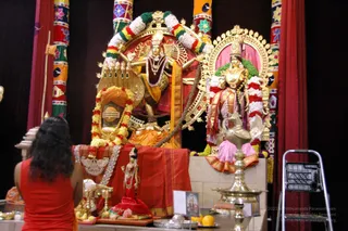 A photograph of a temple shrine with deity murtis dressed in silk and adorned with garlands of flowers in red, white and yellow. A dancing or standing deity form with multiple arms and a crown is at centre, beside a second figure in pink and orange silk with a crown and garland. A brass oil lamp stands at right, and a woman with long dark hair in a red sleeveless top kneels with her back to the camera at lower left. Ornate gold arches, colourful painted pillars and dark red curtains frame the shrine, with a folding metal stepladder at right.