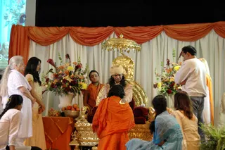 A photograph of a darshan on a stage with orange and cream drapery behind it. A figure with long dark hair, a pale orange turban and a cream-coloured garment sits on an ornate gold throne with a gold canopy, hands pressed together in greeting. Beside the throne a man in orange robes stands, and a kneeling person in orange drapes bows before the seat. Flower arrangements in tall white vases flank the throne, and a table with a fruit bowl and an orange cloth sits at left. Standing visitors include an older woman in white, a young woman in pale yellow, a girl in white, and a man in a white shirt; a seated woman in blue sari sits at lower right. A projected screen shows a blue image at upper left.