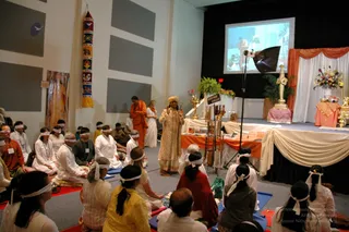 A photograph of a meditation or yoga session in a hall with a stage at the back. A standing man in a cream embroidered kurta and a golden-yellow turban faces a seated group of about 30 people wearing white headbands tied over their eyes; they sit on blue and red mats and cushions. A man in an orange robe stands near the left wall, and a woman in white stands behind the seated rows. On the stage, orange and white draped fabric, a large gold figure, a flower arrangement and a projector screen showing a photo are visible, and a table holds books and ritual items. A tall colourful wall hanging and a portrait of a robed figure hang on the left wall.