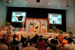 A photograph of a stage event in a hall with a black backdrop and two projection screens showing live imagery. A standing man in a peach and gold embroidered kurta with matching dhoti and a cream turban stands beside a golden throne draped in red, flanked by orange and cream drapes, floral arrangements and potted ferns. Orange-robed attendants stand at the stage edges and a row of seated devotees with white headbands, some in white and orange clothing, face the stage. Tall brass lamps and a golden kalasha-like vessel sit on the stage, with booklets displayed on tables. Spotlights on stands light the scene.