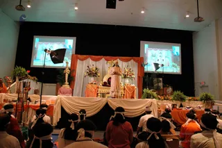 A photograph of a stage event in a hall with a black backdrop, orange and cream drapes, and two projection screens showing a decorated room. A standing figure in a pink and gold embroidered ensemble with a matching cap stands before a gilded throne flanked by floral arrangements and potted ferns. Seated attendees in the foreground wear white headbands and white cloth wraps, several in orange and red clothing. Display tables along the stage hold books and items, and a golden ceremonial sword stands at the left edge. Two spotlights are set on stands to the sides of the stage.