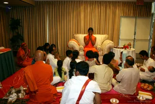 A indoor hall with gold curtains where a seated figure in an orange robe sits on a cream upholstered armchair with clasped hands, facing a seated congregation on red and pink cloths. In the foreground a bald man in an orange robe sits with palms pressed together beside a silver tray with brass vessels. Devotees in white and cream clothes sit on the floor with folded hands, and a child in a white outfit kneels nearby. A small table with a flower arrangement, a kalasha and fruit stands at right, and steel bowls sit on the floor at lower right. Several people appear to be in a seated puja or satsang gathering.