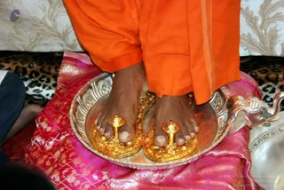A close photograph of bare feet standing on a polished silver tray with a raised embossed rim, resting on a pink and gold brocade cloth. The legs wear saffron-orange dhoti-style cloth. Two ornate gold sandals with small red-jewelled knobs sit on the tray beneath the feet, and a silver elephant-shaped vessel is visible at the right edge against a leopard-print cloth. This appears to be a padapuja-style foot worship ritual, with the feet of a person in orange being honoured.