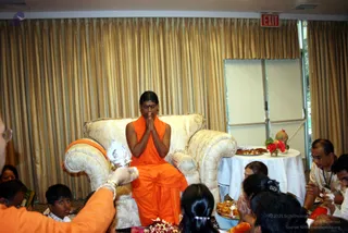 A seated man in an orange-saffron sleeveless robe sits in a cream upholstered armchair with gold-patterned fabric in a hall with gold curtains and a red exit sign. He sits with palms pressed together in namaste, with a dark forehead mark. In the foreground, a man in white on the left holds a silver aarti or puja plate, and seated devotees, including children and women with hair clips, sit on the floor with hands folded. A white-draped table to the right holds a pomegranate, red flowers and a brass tray of offerings. Flower petals lie on the floor by the chair. Whether the seated man is a person cannot be determined from appearance alone, so the identity is left unsure.