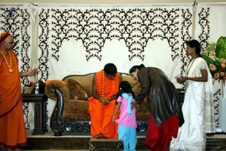 A photograph of a seated figure in an orange robe on a carved wooden sofa with brown upholstery, set before a cream backdrop with dark floral-scroll patterns. The seated man has short dark hair and holds something in his hands, and a garland of beads hangs at his neck. A child in a pink top and light blue trousers with a blue scarf stands before him, while a woman in a dark brown jacket and red skirt bends forward to bless or touch the child. On the left, a man in an orange robe with a gold necklace holds incense sticks, and on the right a woman in a white sari with a dark border stands holding a small plate near a floral arrangement of orange roses and greenery. A small side table holds a brass vessel and fruit.