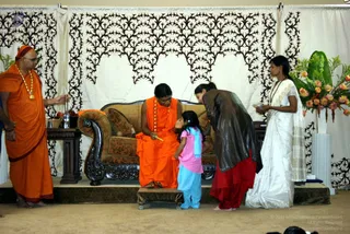 A stage event with a white backdrop decorated in black filigree and a floral arrangement of orange and pink blooms at right. A seated figure in orange robes with an orange-and-beige shawl and a long beaded mala sits on a brown upholstered sofa, holding a yellow object. Before them, a small girl in a pink top and light blue trousers with a blue scarf stands at the sofa's side, while an adult in a dark brown jacket and red skirt bends forward toward her. At left, a standing man in orange robes with a gold necklace and orange head covering holds incense sticks near a small table with a brass vessel and fruit. At right, a woman in a white sari holds a small dish. Several people are visible, including partially seen figures at the edges.