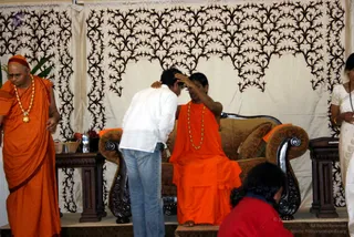 A photograph of a seated figure in a saffron-orange robe and a long orange-and-gold bead necklace on a carved dark wood sofa with cushions in front of a cream backdrop with dark black filigree patterns. A man in a white shirt and jeans bends forward, one hand on the seated figure's forehead, in a blessing gesture. A man in saffron robes with a red headband and a gold pendant necklace stands at the left edge, and a woman in a white sari stands at the right. A person in a red top kneels at the lower right. Side tables hold fruit, a bottle and a basket.