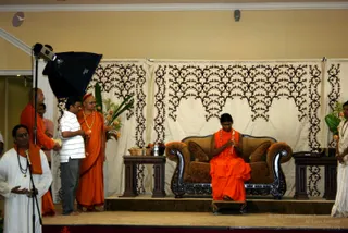 A photograph of an indoor stage event in a hall with cream walls. A seated figure in saffron-orange robes with a long orange garland sits on a dark carved wooden sofa with rust-coloured cushions, holding a rosary-like mala in both hands with eyes lowered. Beside the sofa are small dark wooden side tables with flowers and a bowl. Behind is a cream drape with black scrollwork, and a tall green plant with orange flowers. Standing to the left, a man in an orange shawl holds a small object, while another man in a white shirt holds a mic-like item near a studio light. Further left, a man in white clothing with an orange shawl speaks into a microphone. A figure in white stands at the right edge. A stage with a dark carpet holds the group.