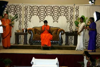 A seated figure in an orange robe and orange shawl sits on an ornate brown sofa with carved black wooden arms on a raised stage before a cream draped backdrop with dark leaf-pattern cutouts. The seated person holds a small instrument or object to their face. To the left, a man in an orange robe with a red head covering and a gold-beaded necklace stands near a wooden side table holding a basket and a flower arrangement. To the right, two women stand: one in a white sari with a beaded necklace and one in a purple sari with a gold border, both beside green foliage and orange flowers. Two young girls in white and gold sit at the lower right edge of the stage, and a white-draped table stands at the front.