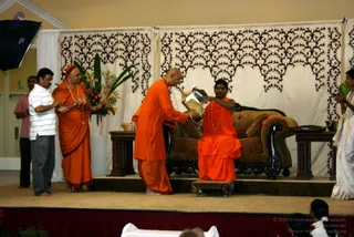 A photograph of a stage event with a long-haired figure in saffron-orange robes and rudraksha mala seated on a carved dark wooden couch with brown upholstery, in front of a cream drape with black leaf-pattern cutwork. A bald man in orange robes stands before him and offers a wrapped gift in yellow paper. To the left, a monk in orange with a red head cloth and mala stands beside a man in a white striped shirt and grey trousers, clapping. At right, a woman in a white sari claps, and a flower arrangement of orange and red blooms and green leaves stands on a side table. Audience members are visible at the bottom edge.