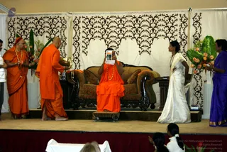 A stage event with an ornate cream curtain patterned in dark brown filigree and tall floral arrangements of orange and pink flowers with large green leaves. A seated figure in full orange-saffron robes and a long orange-beaded mala sits on a dark carved wooden sofa with brown upholstered cushions, holding up a small printed booklet or card with an orange cover so that it covers the face. To the left, a standing man in orange robes and a red headpiece wears a gold-beaded necklace and holds a small ritual item. Another man in orange robes, barefoot and bald, stands beside him holding a gold-toned vessel. On the right, a woman in a white silk sari with a gold border stands near a small wooden side table, and a woman in a violet-blue sari stands at the edge. Two seated audience members in white are visible at the bottom edge of the stage.