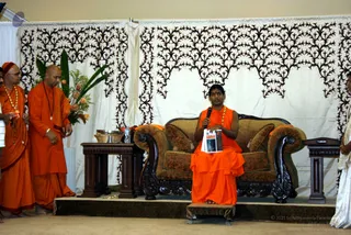 A woman in orange robes sits on an ornate carved wooden sofa with brocade cushions on a low stage, holding a microphone in one hand and a printed booklet in the other. Two men in saffron-orange robes stand to the left, one wearing a rudraksha mala and gold beads, the other holding a small object. A flower arrangement with orange roses and green leaves sits on a side table beside a wicker basket. The backdrop is cream draped fabric with dark black filigree patterns. A person in white stands at the right edge.