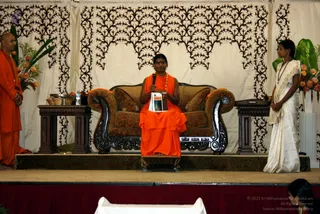 A photograph of a stage event with a central figure seated on a dark carved wooden sofa with brown cushions, wearing an orange saffron robe and an orange beaded necklace, holding a microphone in one hand and a small book in the other while speaking. To the left, a bald man in orange robes stands beside a wooden side table holding a basket and a silver bowl. To the right, a woman in a white sari with a gold border stands beside a wooden table. Behind them hangs a cream backdrop with black ornate scrollwork, flanked by floral arrangements of orange roses and green foliage. A red stage skirt runs along the base, and a white-draped table is visible in the foreground. Three physical people are visible.