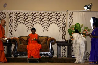 A photograph of a stage event with a seated figure in saffron-orange robes and a long orange-and-gold beaded necklace, holding a small book, seated on an ornate dark carved wooden sofa with brown upholstered cushions and orange trim. Behind the sofa hangs a white curtain with black leaf-scroll patterns. A bald man in orange robes stands at left holding a silver tray or bowl. At right, a woman in a white and gold saree bends forward beside a tall arrangement of green leaves and orange roses, while another woman in a purple saree stands at far right. Wooden side tables flank the sofa and a studio light is mounted at top right. Three physical people are visible plus the seated figure, four in total.