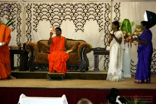 A stage event with a seated man in saffron-orange robes and a rudraksha mala sitting on a brown upholstered sofa with carved dark wood arms, holding up a small card or booklet. To his left, a man in orange robes stands partially at the frame edge holding a bowl. To the right, two women stand: one in a white sari with gold border holding her hands together, and one in a purple sari with gold border holding a paper. Behind them are a potted tropical plant and orange flower arrangement, and a black-and-white patterned drape backdrop. Foreground audience heads are visible at the bottom.