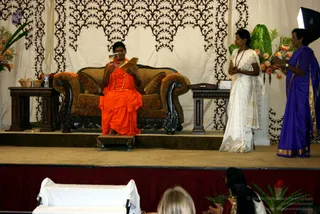 A photograph of a stage event with an ornate carved wooden sofa upholstered in brown and gold fabric at centre. A figure in a long saffron-orange robe with a rudraksha mala and red garland sits on the sofa, holding up a small card or photograph in both hands. To the right, a woman in a white saree with gold border stands with hands folded in front of her, beside a woman in a purple saree with gold border who gestures with one hand. Behind them are a cream backdrop with black filigree patterns, tall floral arrangements with orange blooms and green leaves, and a small side table with a box. In the foreground lower edge, a blonde-haired person and a woman with a long braid in a red top are seen from behind, facing the stage, with a white-draped lectern at left.