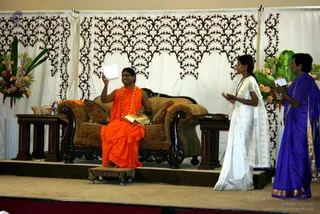 A photograph of a stage event with a draped cream backdrop patterned in dark brown scrollwork. A man in an orange saffron robe and orange-red garland sits on an ornate carved wooden sofa with brown upholstery, one hand raised holding a white card and the other resting on a gold-coloured object, his feet bare on a small footstool. To the right, a woman in a white sari stands with palms pressed together, and beside her a woman in a purple sari with gold border holds a small white card. Floral arrangements of orange flowers and green leaves flank the stage, and small wooden side tables sit beside the sofa. The man is a person by appearance: long swept-back hair and saffron robes with a rudraksha-style garland, though appearance is only partly clear.