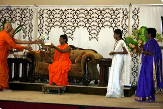 Photograph of a stage event with a long-haired figure in saffron-orange robes and an orange bead garland seated on a brown upholstered sofa with carved dark wooden arms, a small wooden footstool before him. He holds up a book or framed item that a bald monk in orange robes on the left reaches toward. A woman in a white saree with gold border stands at right beside a woman in a purple saree with gold border who holds a large arrangement of orange flowers and green leaves. Behind them hangs a cream backdrop with dark brown leaf-vine patterning. Hidden by the visible cues alone, the seated figure's identity is uncertain, so no candidate is asserted.