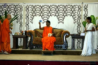 A stage with a white backdrop patterned in dark brown filigree and tropical leaves at the sides. A seated man in orange robes and a rudraksha mala holds up a small printed photo card while smiling, seated on an ornate carved brown upholstered sofa. To the left, a bald man in orange robes stands beside a table with a basket of fruit and flowers. To the right, a woman in a white sari with a dark shawl stands near a small side table with books. A low stool sits in front of the sofa. Three people are visible.
