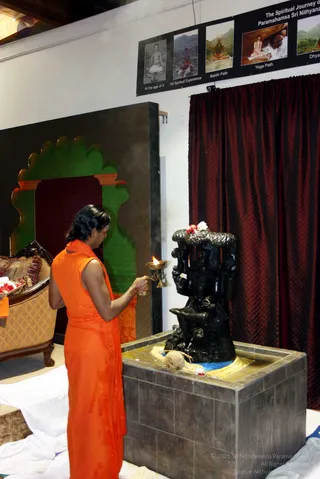 A man in a sleeveless saffron-orange garment stands at a tiled stone pedestal, holding a brass oil lamp with a lit flame toward a dark stone deity murti with multiple arms and a flower-garlanded crown. A coconut, yellow cloth and flower petals sit on the pedestal. Behind him, a green arched panel frames an ornate upholstered chair with flower offerings, and a dark red draped curtain hangs on the right. Above, a black banner shows photographs of a spiritual journey labelled At the age of 9, 1st Spiritual Experience, Bakthi Path, Yoga Path and Dhyana, with a caption at top reading The Spiritual Journey of Paramahamsa Sri Nithyananda. The scene suggests a puja or aarti at a ceremonial display.