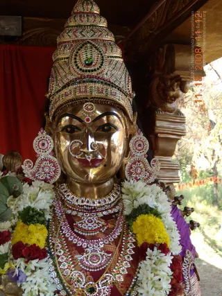 Close photo of a gilded metal deity murti. It wears a nose ring, jewelled earrings, layered gem-studded necklaces and a red-and-gold garment, and is garlanded with white jasmine and yellow and red flowers. A red cloth backdrop and carved wooden architecture frame the image, with greenery and a dated timestamp at the edge.