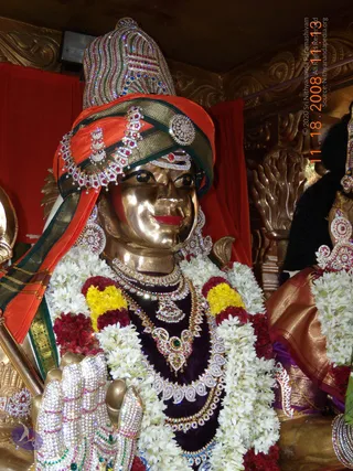 A golden metal statue bust of a figure wearing an orange and green turban decorated with red and white stones and a jewelled crest, with a golden face, heavy jewelled necklaces, earrings and a garland of white jasmine and yellow and red flowers. The jewelled hand raised in a gesture on the left is also studded with stones. Behind it hang red drapes and ornate gold carvings. The statue is the only figure shown; no living people are visible.