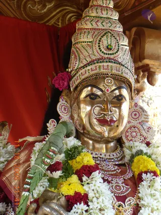 Close-up photograph of a gilded metal deity murti. The face has large almond eyes, a red bindi, a nose ring set with stones, and a red-lipped smile. Heavy jewelled earrings, a multi-strand jewelled necklace and a pink rose in the hair adorn the figure. Garlands of white jasmine and yellow and red flowers drape the shoulders, with a green leaf bundle at the left. A red cloth backdrop and carved gold woodwork frame the scene. The image is an ornamental decoration for a procession or ritual display.
