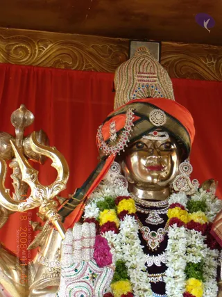 A gilded metal murti of a deity with a bronze-gold face, wearing an orange and dark green turban with a jewelled crest and a gem-set brooch. Ornate paisley-patterned earrings, layered jewelled necklaces and a rudraksha-style ornament adorn the figure. A large sparkling silver-and-jewel hand, palm outward, extends toward the viewer. Strands of white jasmine, yellow marigold and red rose garlands drape the shoulders. Beside the figure a gold trident and a long-stemmed gold instrument are visible against a red draped backdrop, with ornate gold carved wall panels above. No people are present; the figure is a statue.