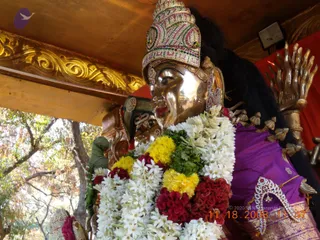 A close photograph of a gleaming metal deity murti on a processional setting, its face polished bright gold-bronze and wearing a jeweled crown with green, pink and white stones. It is draped in a purple silk garment with a gold-bordered pattern and adorned with a wide garland of white jasmine flowers, yellow marigolds, green leaves and red celosia blooms. A golden ornamental arch and a gilded hand-shaped element stand at the right, and trees are visible through an open side. A date stamp in red digits and a copyright line appear in the lower corner.