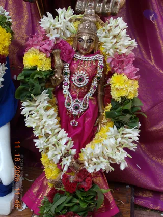 A photograph of a bronze deity murti standing in front of a magenta and gold brocade backdrop. The figure wears a magenta silk garment with gold zari borders and a jewelled necklace set with white stones, green and pink stones. It holds a small object in one hand, and its head wears a crown. White jasmine garlands, yellow marigolds, pink flowers, green leaves and red celosia surround the figure. A partial white statue and blue fabric appear at the left edge, and the lower right shows scattered petals on a wooden surface.