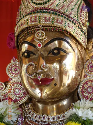 Close-up photograph of a gleaming gold metal deity murti face adorned with a jeweled crown set with white, green and magenta stones, a red-and-white stone forehead ornament, a jeweled nose ring and nose pin, and large jeweled earrings in pink and white. Jasmine-style white flower garlands and a pink flower frame the lower edge, with a red backdrop behind. No people are visible.