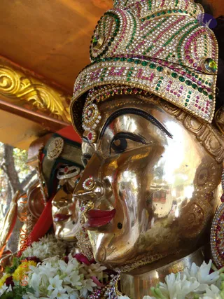 Close-up photograph of a gilded metal deity murti with a polished golden face, dark arched eyebrows, a red-lipped mouth and a small nose ornament. Its jeweled crown is studded with green, pink and white stones, with matching earrings. White jasmine flowers and marigold garlands fill the lower foreground. Behind it, a second gilded face and a red fabric drape are visible under a golden canopy edge. No people are visible.