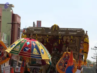 A street procession photographed in daylight, with a decorated ratha (chariot) carrying a carved wooden shrine. A multicolored ceremonial umbrella with red, green, yellow and blue panels, bearing bird and floral motifs and fringed tassels, shelters the front. Yellow and white marigold garlands hang across the shrine, and a dark deity murti decorated with garlands sits within it. Orange flags and decorated banners flank the scene, and a small framed portrait is visible at lower left. Buildings and a utility pole line the street behind. No people are clearly visible.