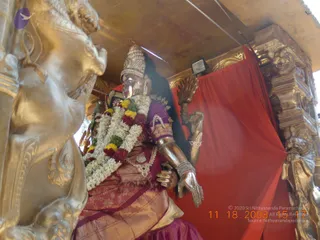 A photograph of a golden ratha chariot shrine housing a metal deity murti seated on a golden bull-shaped vehicle. The murti wears a purple and maroon silk sari with gold border, a tall ornate crown, a long garland of white jasmine, yellow and red flowers, and gold jewellery including a large necklace and bangles. A red cloth hangs behind it, and gilded carved lion-faced pillars frame the shrine. No physical people are visible.