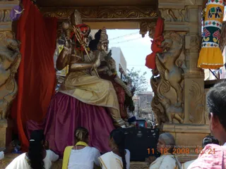 A gilded deity murti with a trident is seated on an ornate golden ratha (chariot) decorated with carved lions and elephant reliefs and draped with red and magenta cloth. The murti wears a gold garment, bangles and a garland of white and orange flowers. Below, several women in white saris and a girl in a yellow top sit on the chariot platform beside a black amplifier. Street buildings and a tree are visible through the open side of the ratha during a procession. A colourful painted cylinder hangs at the right edge.