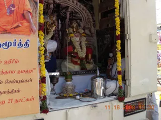 A photograph of a decorated shrine built into the side of a white vehicle, with a golden deity murti in a red and gold garment, adorned with white and yellow flower garlands and red blossoms. Behind the murti is a painted backdrop of a temple arch and a banyan-like tree. In front sit a small brass oil lamp, a kalasha-like stainless steel pot, a brass vessel, a leaf-topped bowl and a plate of flowers. On the left, a large poster shows a saffron-robed seated figure with rudraksha beads, above Tamil script. A man in white ritual clothes holds a microphone at the right of the shrine, and a second man in white sits partly visible behind the deity.