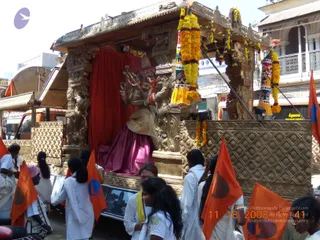A golden ornate ratha (chariot) with carved pillars, red drapes and a seated deity murti decorated with marigold garlands, moving through a city street during a procession. Devotees in white clothing and some in yellow walk alongside, several holding orange flags with a blue and white emblem. Shops and multi-storey buildings line the road. Dated 11.18.2008 in the corner of the photo.