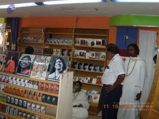 Interior of a book and merchandise shop with orange and green painted walls and wooden shelves stocked with books and framed photographs. On the left, a wall of small booklets and cards faces out, and a large black-and-white portrait labelled "PARAMAHANSA NITHYANANDA" hangs on the shelf. A man in a white short-sleeved shirt and dark trousers stands at the right with his hands on his hips, facing the shelves. Beside him, a man in a long white garment with a white shawl and a mala of beads looks toward the books. A woman in white sits on the floor at the lower centre, partly hidden by the shelving. A time and date stamp appears in the lower right corner.