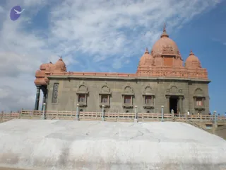 A photograph of a large stone memorial building with a reddish-pink domed roof and several smaller domes, set on a whitewashed raised platform under a partly cloudy blue sky. The grey stone walls have arched recessed windows with barred grilles, a pillared entrance porch on the left, and a central doorway. A low railing runs along the platform edge with small lamp posts. A few small distant figures stand near the entrance.