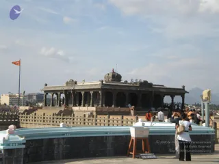 A photograph of a dark stone temple pavilion with a domed roof and a row of carved pillars, set on a raised platform beside a light blue parapet wall. An orange flag flies on a tall pole at the left. Several visitors stand and walk around the platform, including a woman in a white top carrying a beige handbag, and a figure in orange clothing on the steps. A small wooden stand with a sign reading 'Ekaasana Mandapam' rests near the parapet, under a pale blue sky with distant hills.